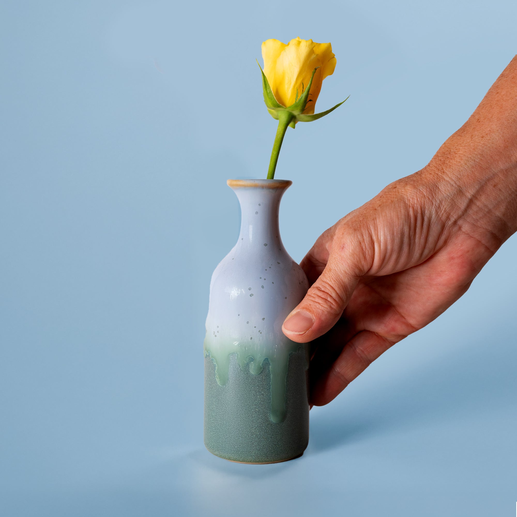 Small handmade flower vase in green and white glaze held by hand with a yellow rose from Pottery For The Planet