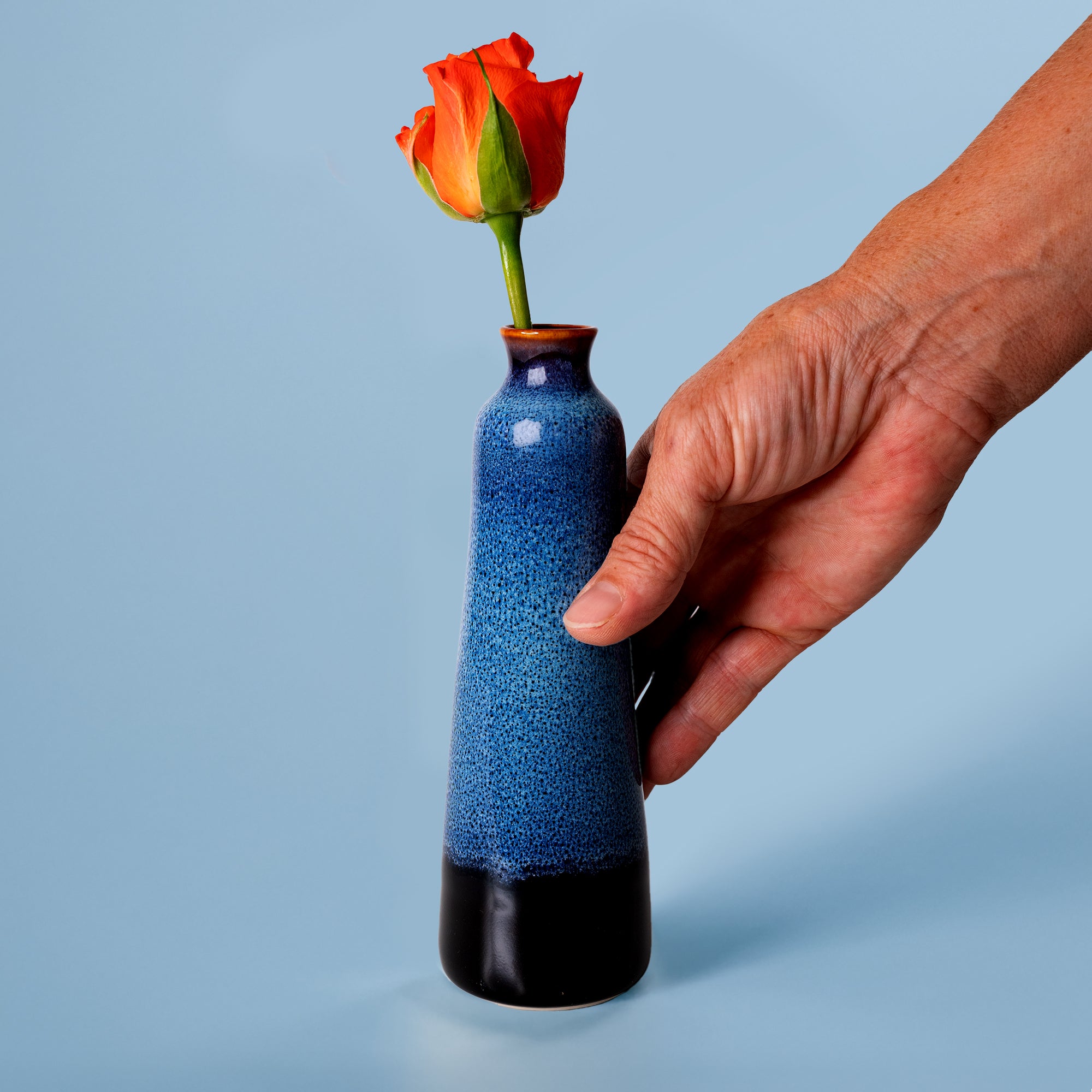 Unique handmade ceramic vase in blue and black held by hand with an orange rose inside