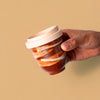 Hand holding artisan pottery travel cup with brown and white glaze and beige lid by Pottery For The Planet