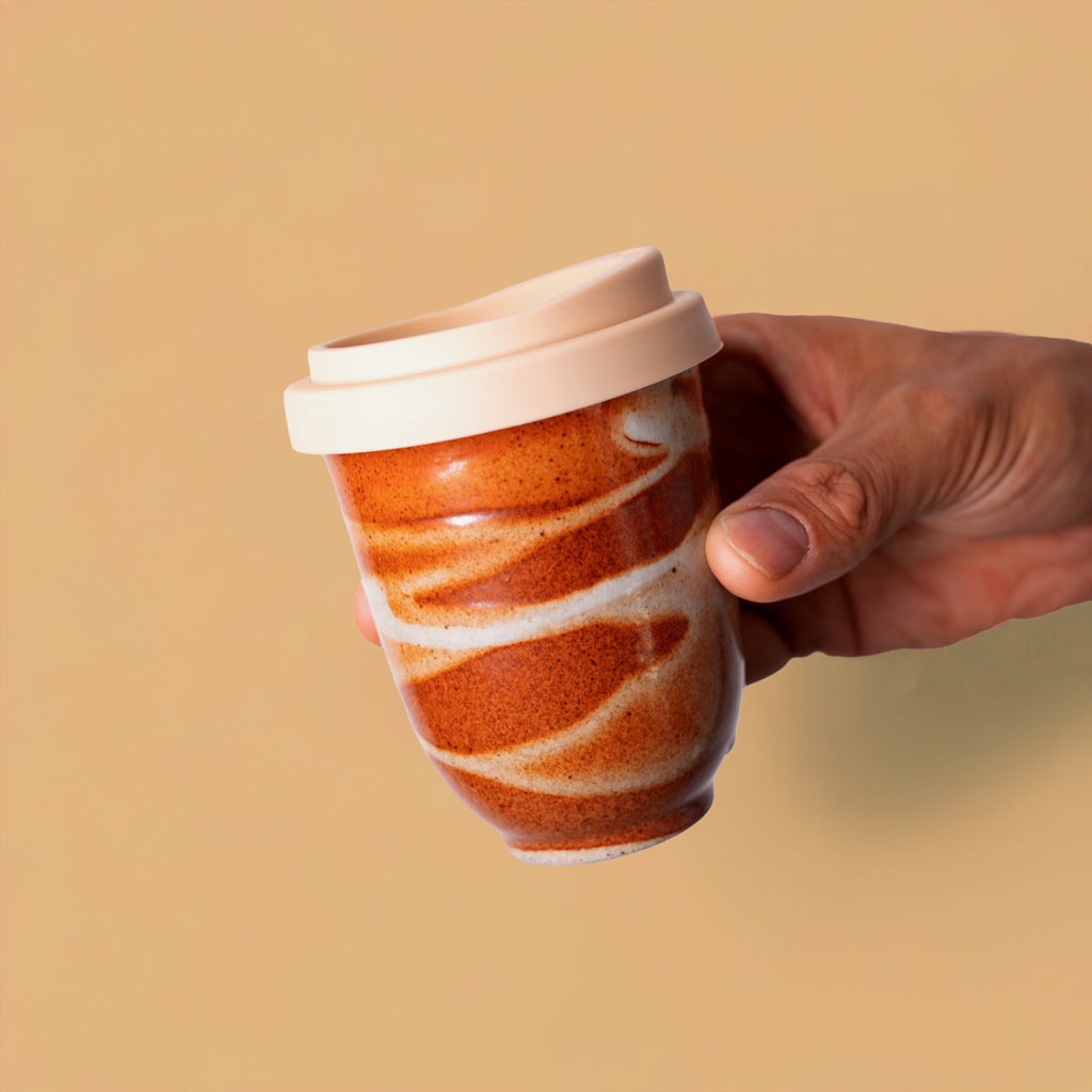Hand holding artisan pottery travel cup with brown and white swirl design and silicone lid
