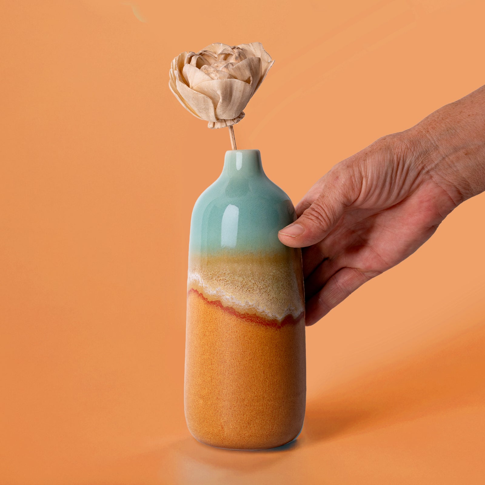 Hand holding a Pottery For The Planet eco-friendly home decor vase with blue and orange glaze and dried flower