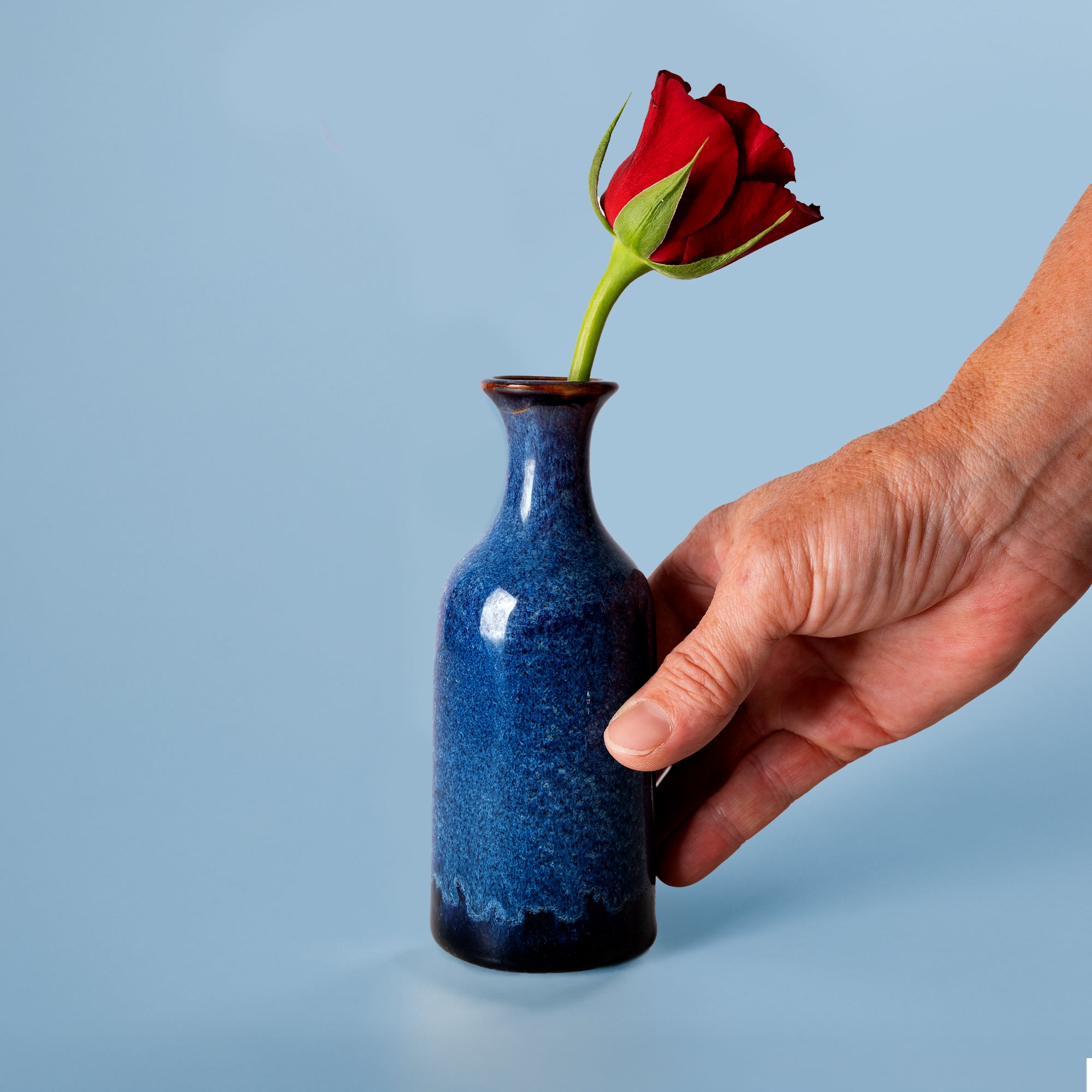 Small handmade flower vase in blue ceramic with a single red rose held by hand