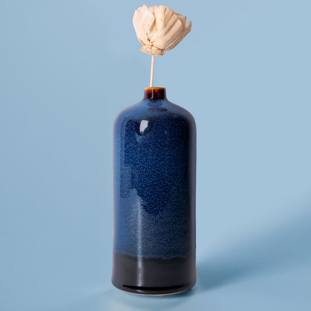 Pottery For The Planet medium decorative bud vase in blue with black base and a dried flower on light blue background
