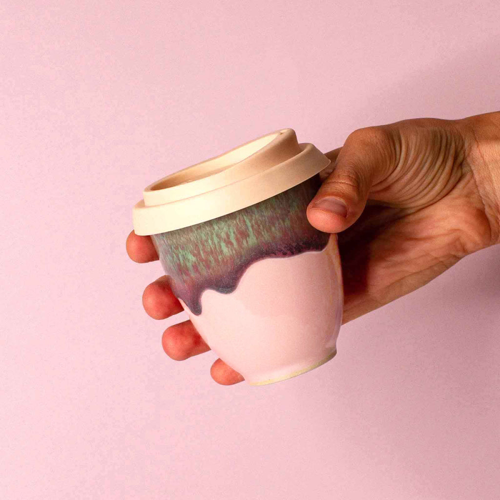 Hand holding a pink ceramic travel cup with green and purple glaze and a beige lid by Pottery For The Planet