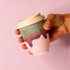 Hand holding Pottery For The Planet pink ceramic travel cup with beige lid and artistic glaze design