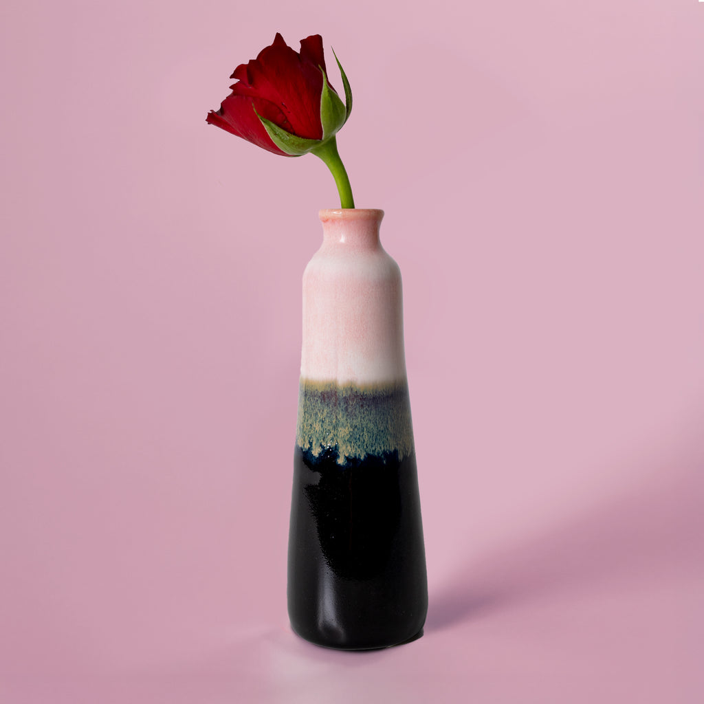 Unique handmade ceramic vase with pink, green, and black glaze holding a red flower on pink background