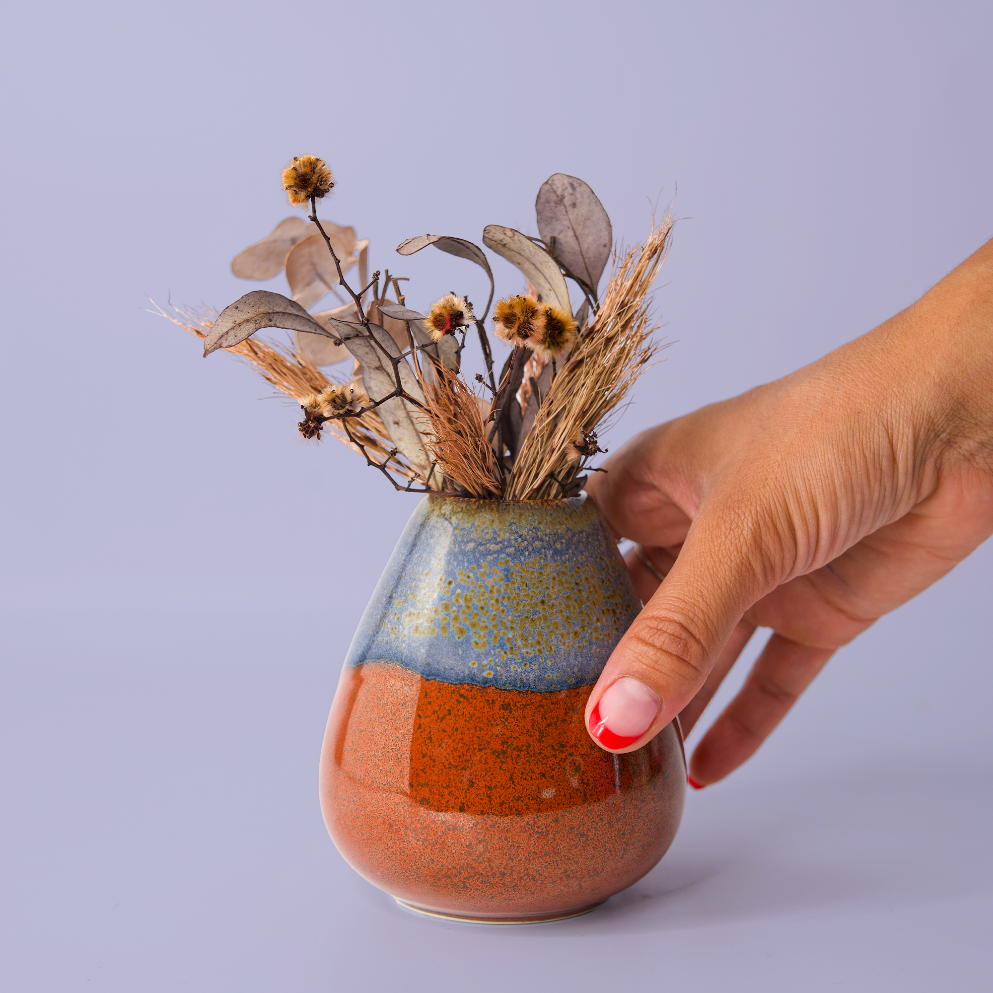 Hand holding a unique pottery flower vase with blue and orange glaze filled with dried flowers