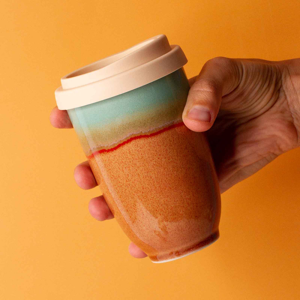 Hand holding a Pottery For The Planet handmade reusable cup with blue and brown gradient design and white lid
