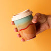 Hand holding a colorful handmade reusable cup with a beige lid against an orange background